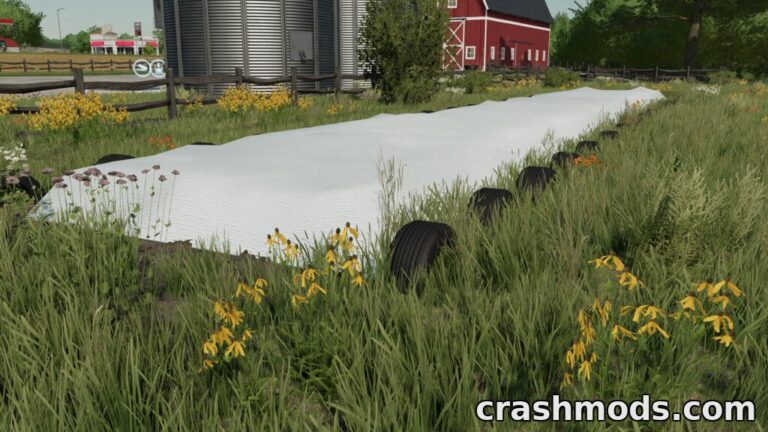 A silage silo covered with white tarp sits in a grassy field, surrounded by wildflowers and farm structures.